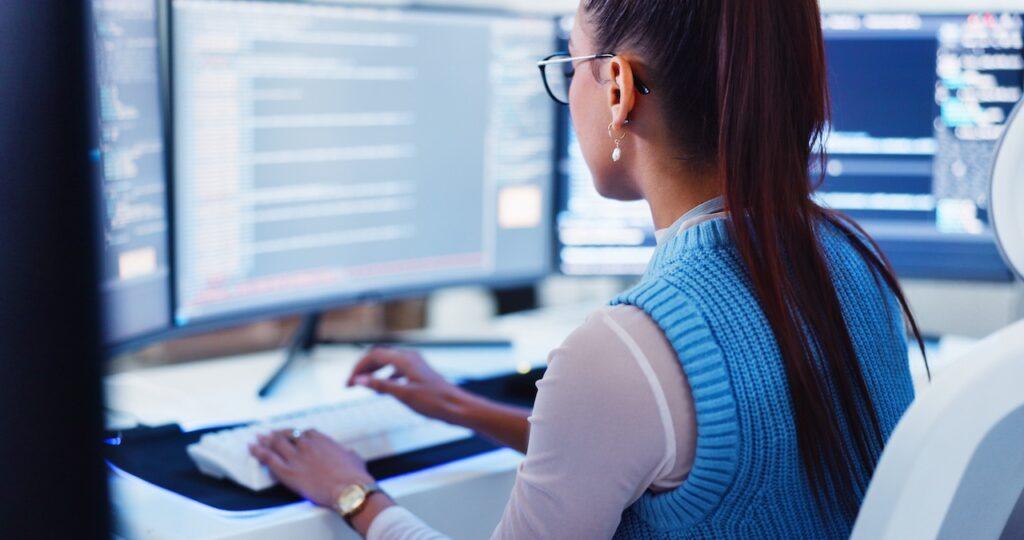 Frau mit Brille arbeitet konzentriert an mehreren Bildschirmen mit Code im Entwicklerbuero
