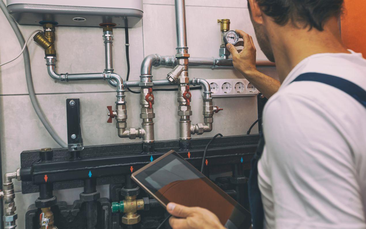 Installateur prüft den Druck einer Hauswasserwerkpumpe mit Manometer und Tablet in modernem Technikraum mit Rohrleitungen