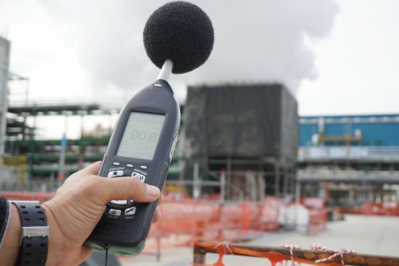Hand hält ein Schallpegelmessgerät mit 90.8 dB vor Industrieanlage – Grundlage zur Planung von Kulissenschalldaempfern im Außenbereich